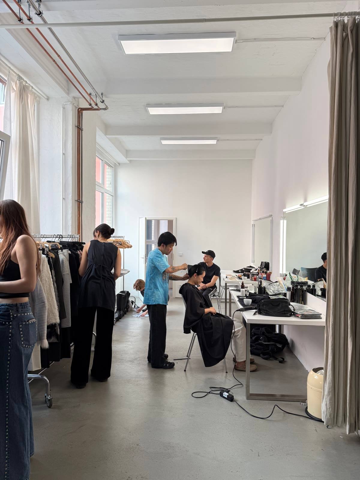 Hair and make-up underway in a bright studio, rails of looks along the wall